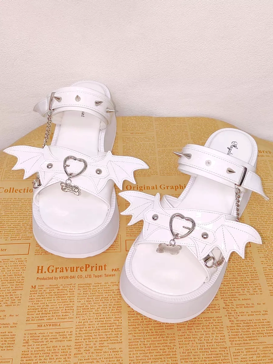 Full front flat lay view of white Gothic Lolita platform sandals showing thick platform sole, ornamental spiked straps, bat wing decorations, heart-shaped buckles, and complete construction detail on newspaper tabletop
