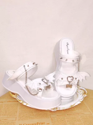 Side oblique view of white Gothic Lolita sandals on retro newspaper tray featuring thick 6.5cm platform sole, spiked ankle straps, and bone-shaped metal pendants, showcasing the commanding height and punk-inspired embellishments