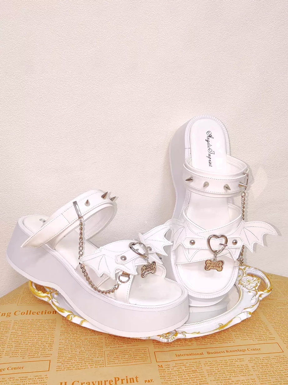 Side oblique view of white Gothic Lolita sandals on retro newspaper tray featuring thick 6.5cm platform sole, spiked ankle straps, and bone-shaped metal pendants, showcasing the commanding height and punk-inspired embellishments
