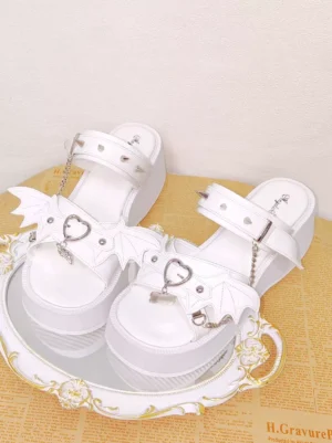 White Gothic Lolita platform sandals with spike rivets and bat wing charms displayed on ornate gilded tray with newspaper background, showing oblique overhead view of glossy white PU leather, heart-shaped metal buckles, and chain pendants