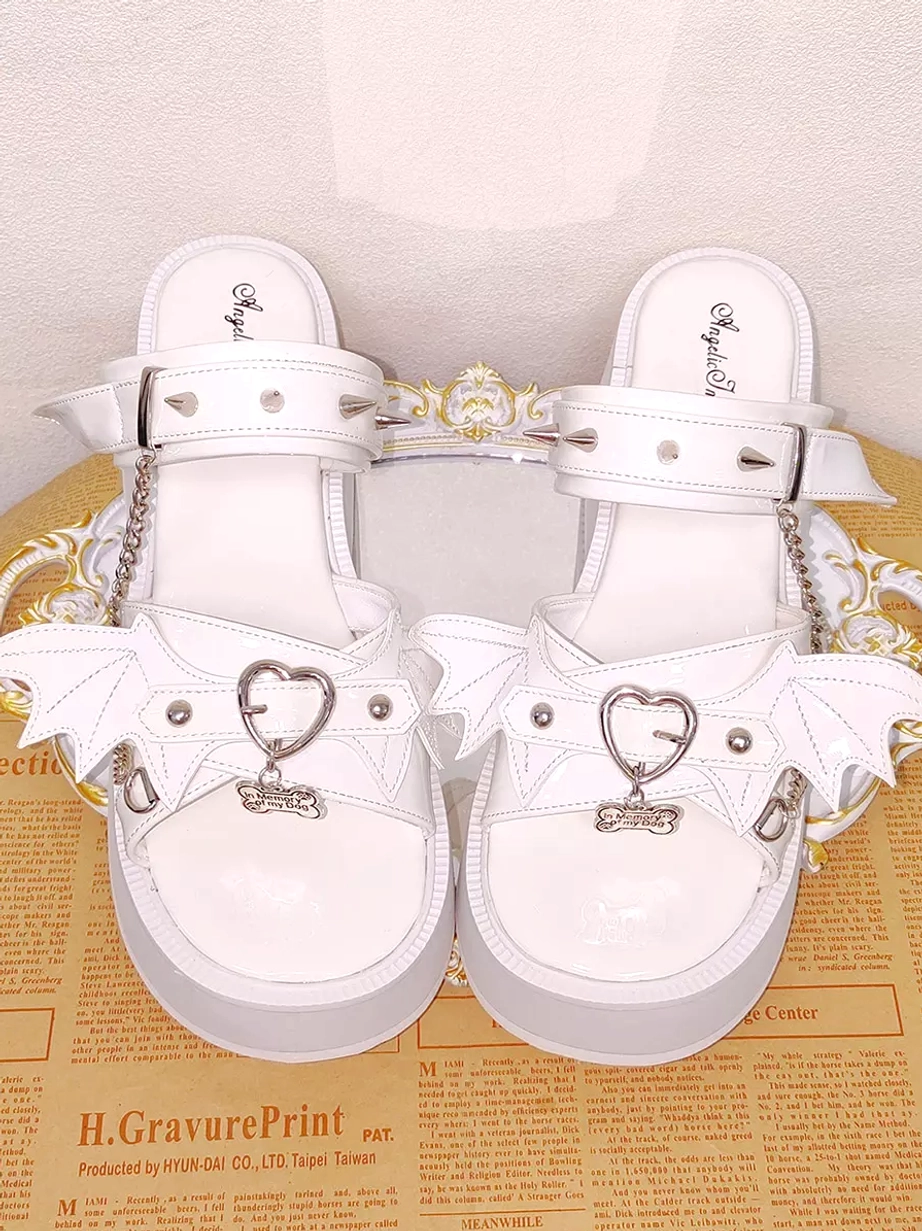 Top-down view of white Gothic Lolita platform sandals on carved vintage tray showing glossy white patent leather finish, bat wing decorations, heart-shaped buckles, and full spiked ankle strap design