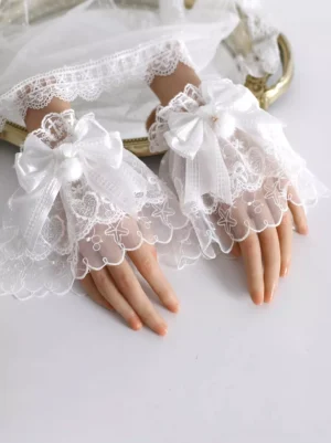 White lace fingerless gloves with satin bows and pom-poms displayed on mannequin hands with decorative gold tray, sweet lolita fashion accessory side view
