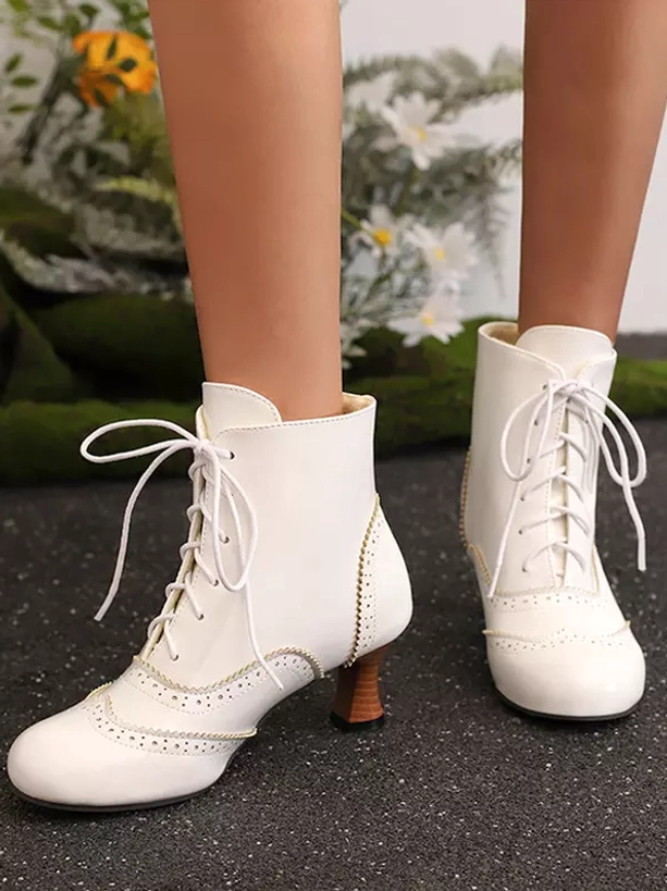White lace-up Lolita ankle boots frontal flat lay view showing smooth PU leather upper, round toe silhouette, and delicate brogue trim details on gravel ground with floral plants