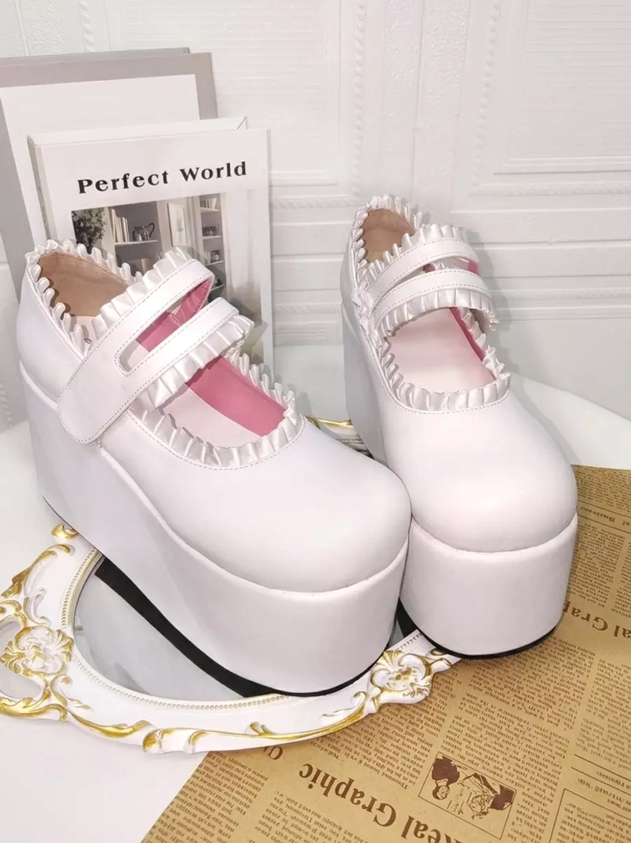 Pair of white lolita platform wedge shoes side three-quarter view featuring velcro ankle straps, delicate ruffled collar, thick white platform sole, displayed with vintage mirror and newspaper styling