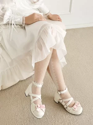 White Lolita chunky heel pumps with pink bow accents and cross strap buckles worn with sheer polka dot stockings, low-angle full leg shot on wooden floor