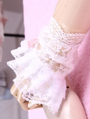 Side close-up view of white layered ruffled lace lolita wrist cuffs on wooden mannequin arm with subtle pink bow accent on pink plush background