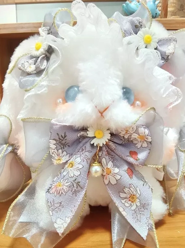 White fluffy plush bunny crossbody bag with blue glass eyes, daisy print bow, and ruffled lace trim displayed on wooden shelf background