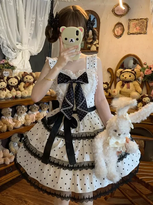White polka dot sweet lolita JSK dress front three-quarter view with black chest bow and tiered ruffle skirt on model with twin buns