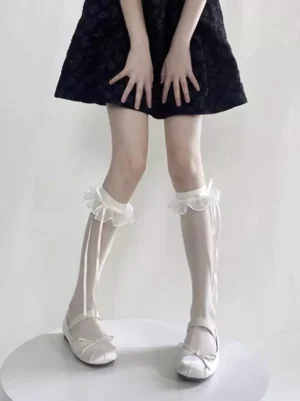 Front full view of white ruffled ribbon calf socks featuring delicate sheer fabric, frilled edge trim, and extended satin ribbon ties on standing model in black mini dress