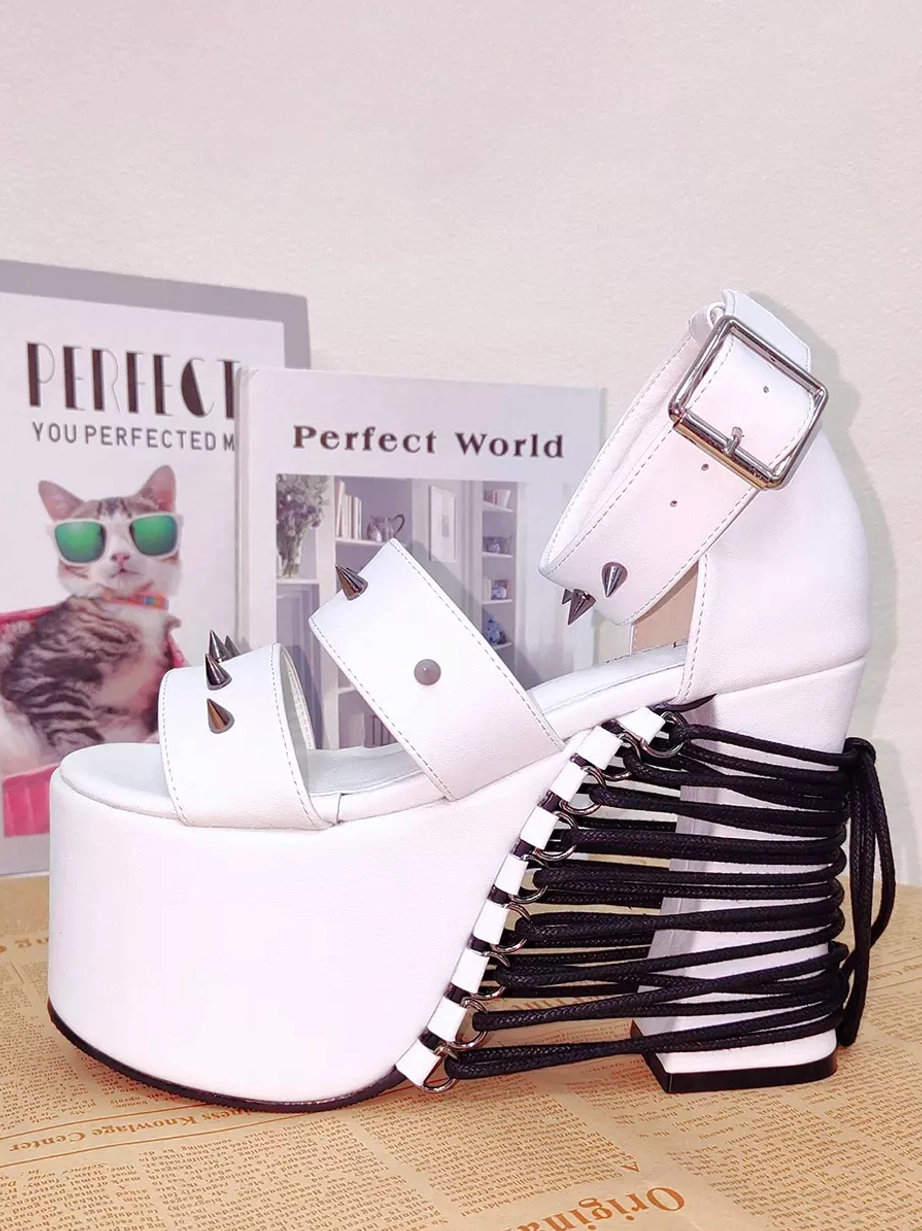 White studded Lolita platform sandals three-quarter side flat lay profile view displaying black lace-up back heel, ankle buckle detail, and rivet embellishments on newspaper backdrop