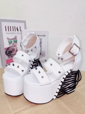 White studded platform Lolita sandals overhead flat lay view with double peep-toe straps, silver rivets, ankle buckle strap, and black lace-up heel detail on newspaper backdrop with cat-themed posters