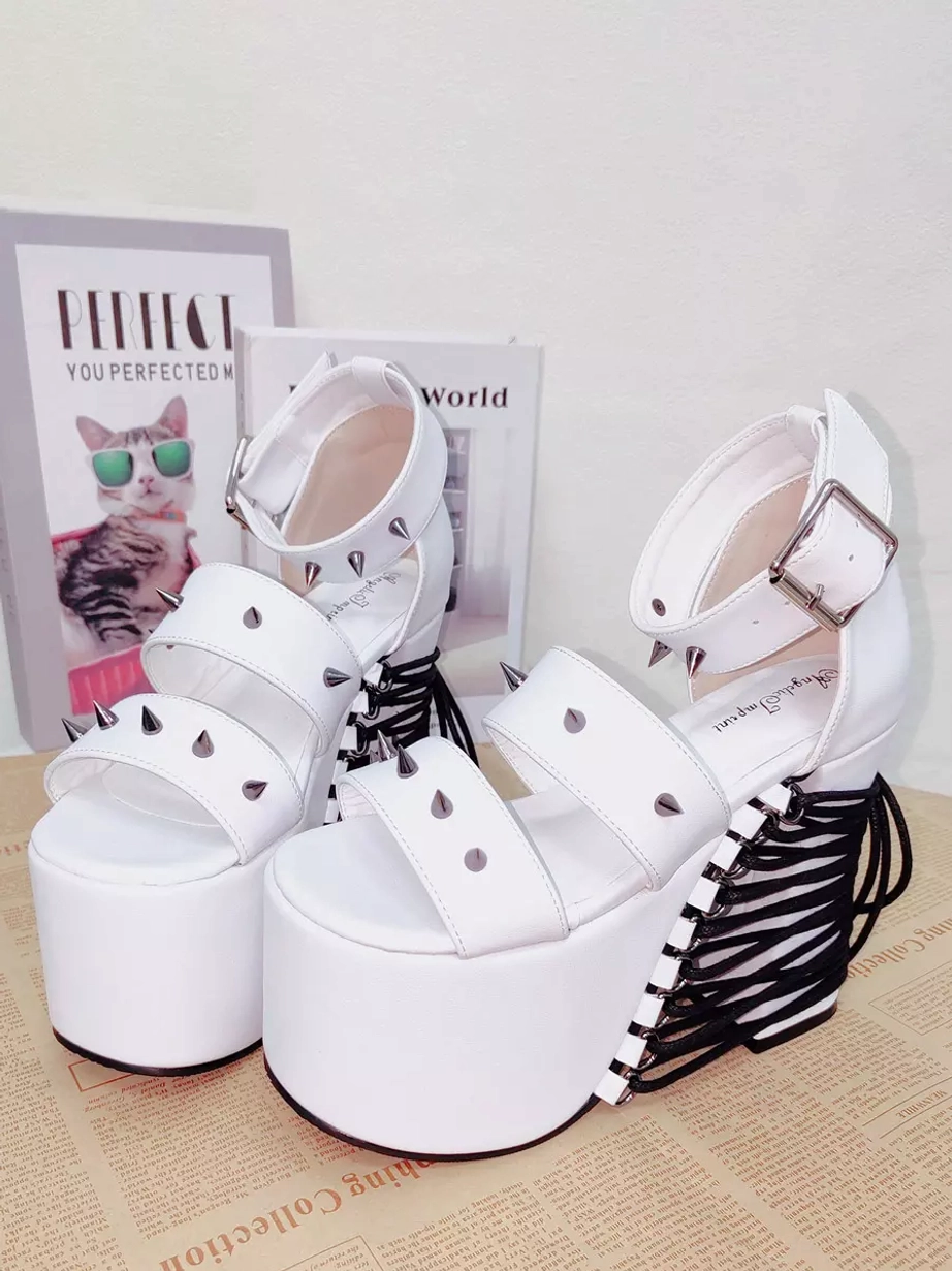 White studded platform Lolita sandals overhead flat lay view with double peep-toe straps, silver rivets, ankle buckle strap, and black lace-up heel detail on newspaper backdrop with cat-themed posters