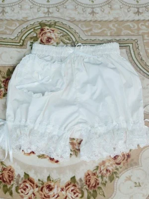 White lolita bloomer shorts with lace ruffled hems and heart-shaped satin bow pocket detail, displayed flat lay on vintage floral carpet, showing adjustable drawstring waist and loose leg silhouette, authentic sweet lolita foundation garment