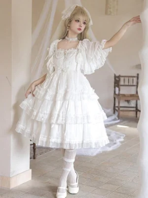 White sweet lolita jumper dress with tiered ruffled skirt, puff sleeves, and pearl accents worn by model with blonde hair, veil and choker in vintage indoor setting