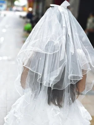 Delicate white layered bridal veil with wavy edge tulle and satin bow top detail featuring pearl embellishments, back view outdoor street setting, ethereal lolita wedding accessory