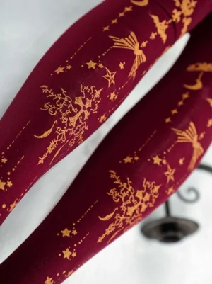 Close-up side angle of wine red velvet lolita stockings showing intricate gold celestial and cross pattern details with vintage decor props